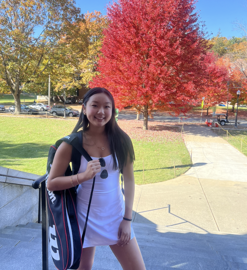 Madison standing on the steps of Alumni Gymnasium holding a tennis racket. There is colorful foliage in the background.