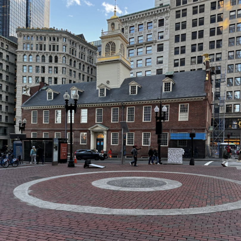 Image featuring Boston's original town hall. The building can be seen from a plaza filled with red bricks. Several buildings can be seen behind the town hall.