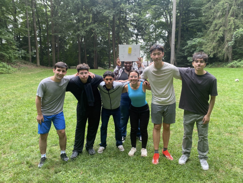 7 students posing on a lush green field surrounded by trees
