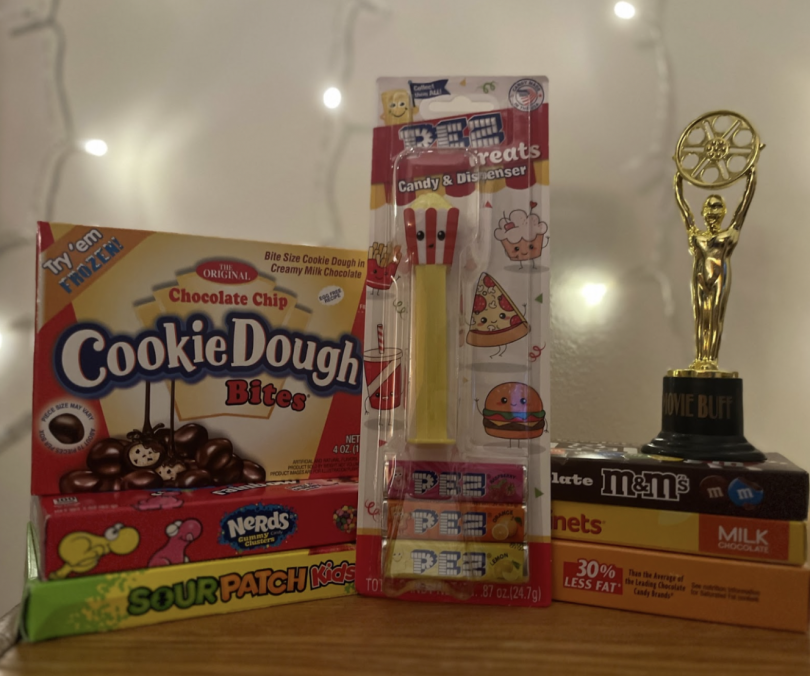 Six boxes of candy are displayed on a wooden shelf. The boxes say "Cookie Dough Bite," "Nerds Gummy Clusters," "Sour Patch Kids," "M&M's," "Raisinets," and "Milk Duds." In the middle of the boxes, a packaged PEZ dispenser, looking like a popcorn bucket, s