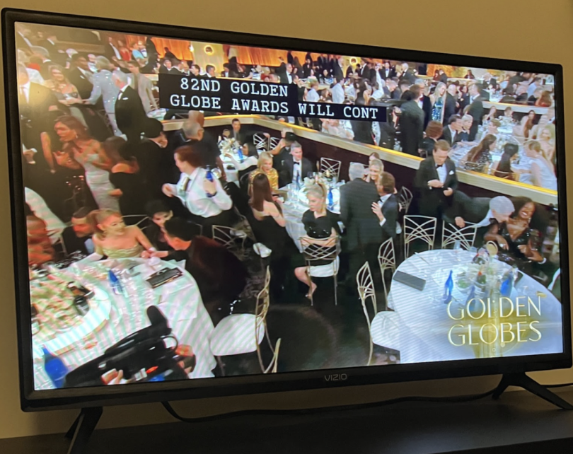 A TV, resting on a wooden stand, displays live footage of a ballroom filled with circular tables, where countless people sit and talk. The screen says "Golden Globes" and the closed captions say "82nd Golden Globe Awards will continue".