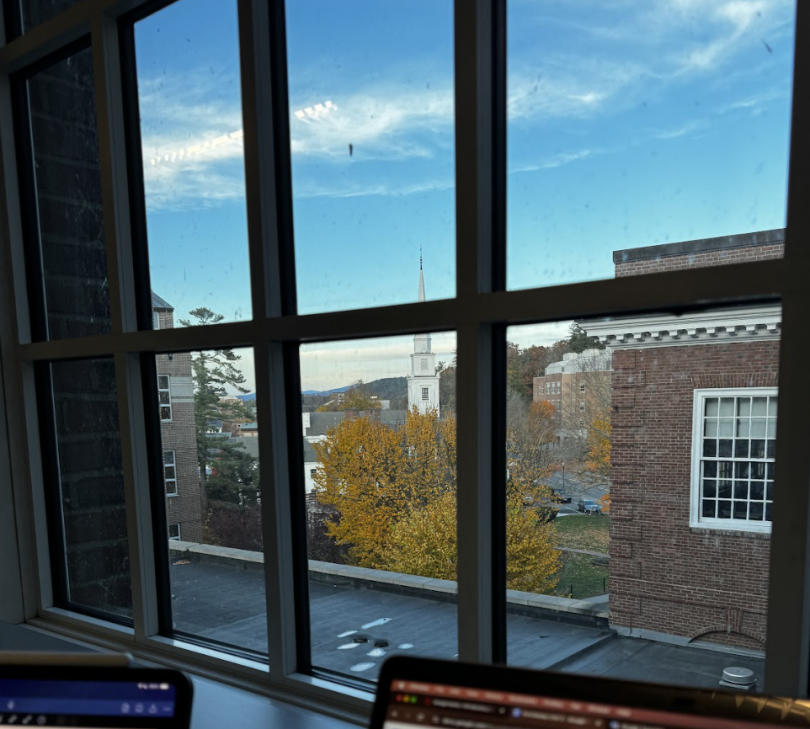 Image featuring the view from outside one of the windows in the stacks at Dartmouth. The image features a desk with a computer and an iPad inside the building while also featuring several trees and buildings outside the window.