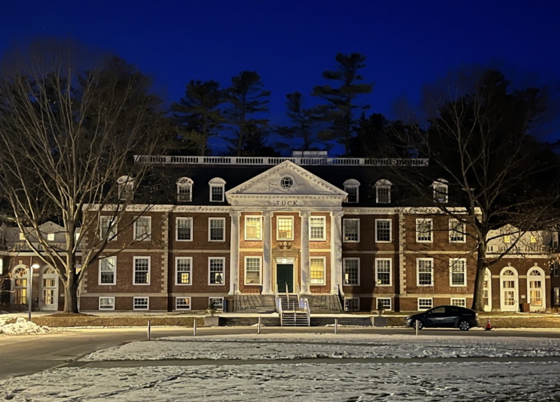 A large brown building with white columns and the word "Tuck" on its roof sits against the night sky and a snowy ground.