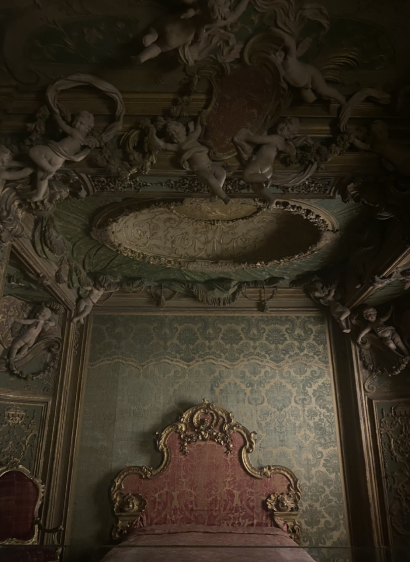 A fancy antique bed with a gold-trimmed headboard, rich fabric, and a ceiling covered in cherubs and decorative carvings.