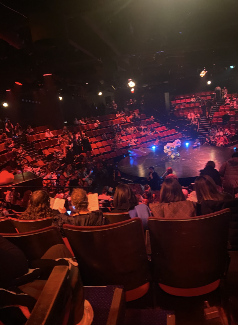 A dimly lit theater with red seating arranged in a circular layout around a central stage. A few members of the audience are seated.