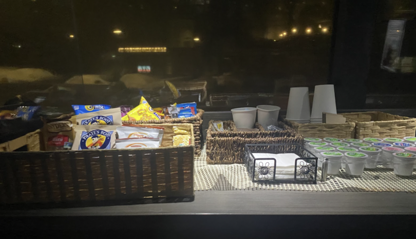Snack and beverage station with wicker baskets holding chips, crackers, and coffee supplies, set against a dimly lit window.