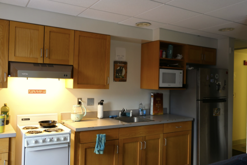 A small kitchen with wooden cabinets, a stove, a microwave, and a fridge with magnets. The countertop holds a kettle, dish soap, a cereal box, and a water bottle.