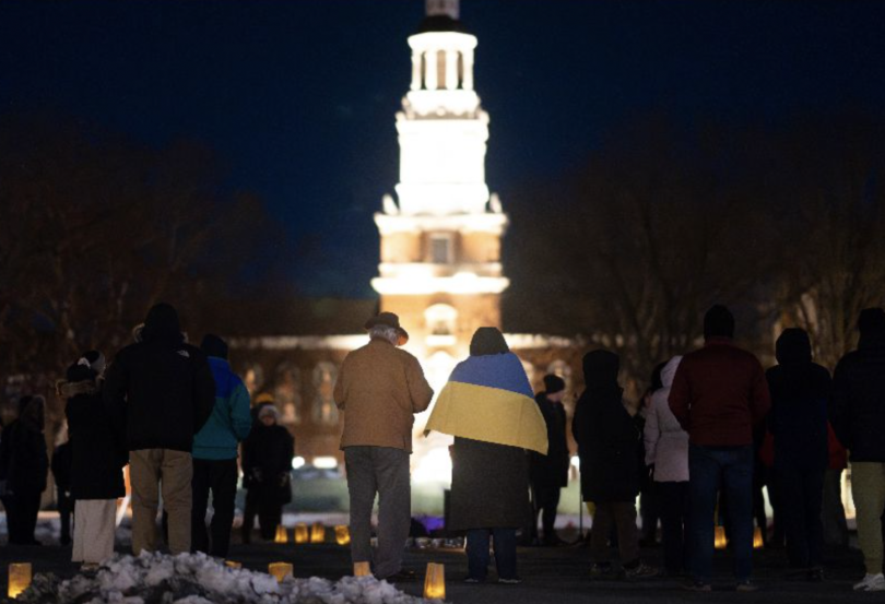 A group of people is gathered outside at night, some holding candles, with one person draped in a Ukrainian flag. In the background, the Baker Memorial Library tower at Dartmouth stands brightly illuminated against the dark sky.