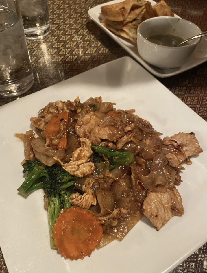 A plate of stir-fried noodles with chicken, broccoli, and carrots. In the background, there are glasses of water, a bowl of soup, and a plate of crispy folded pastries.