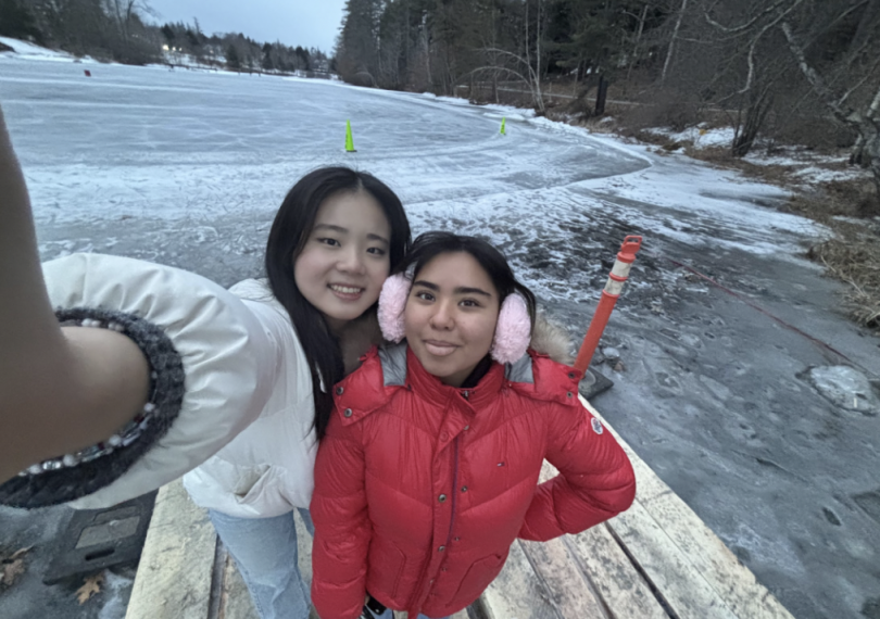 Two young women pose for a selfie on a wooden platform by a frozen lake, surrounded by a winter landscape. One wears a white jacket, and the other a red coat with pink earmuffs.