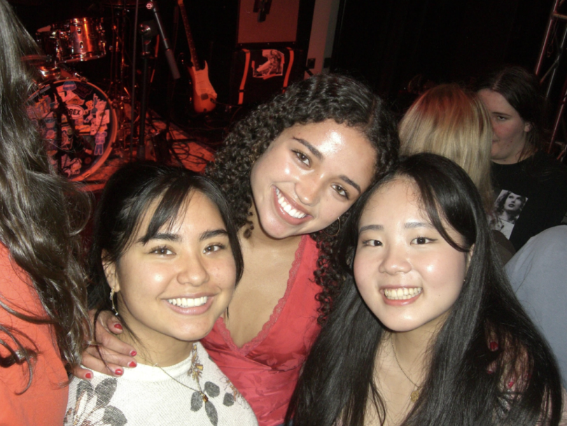 Three young women smiling and posing together at a concert, with a stage featuring musical instruments and a drum set in the background