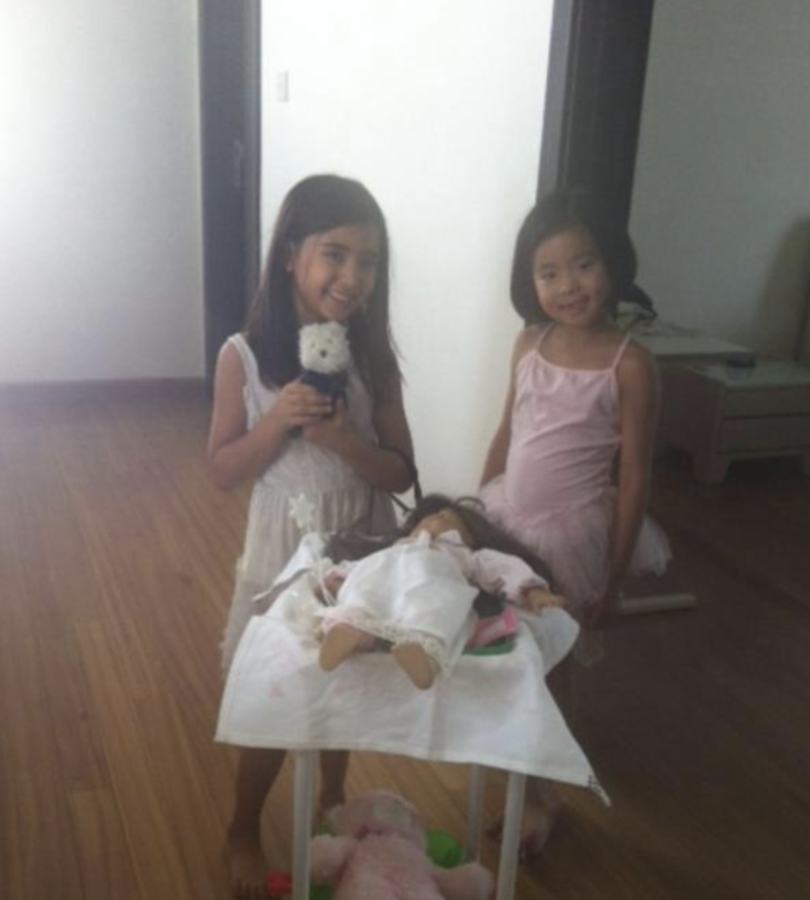 Two young girls smile as they pose beside a small cart. One girl holds a stuffed animal, while the cart carries a doll