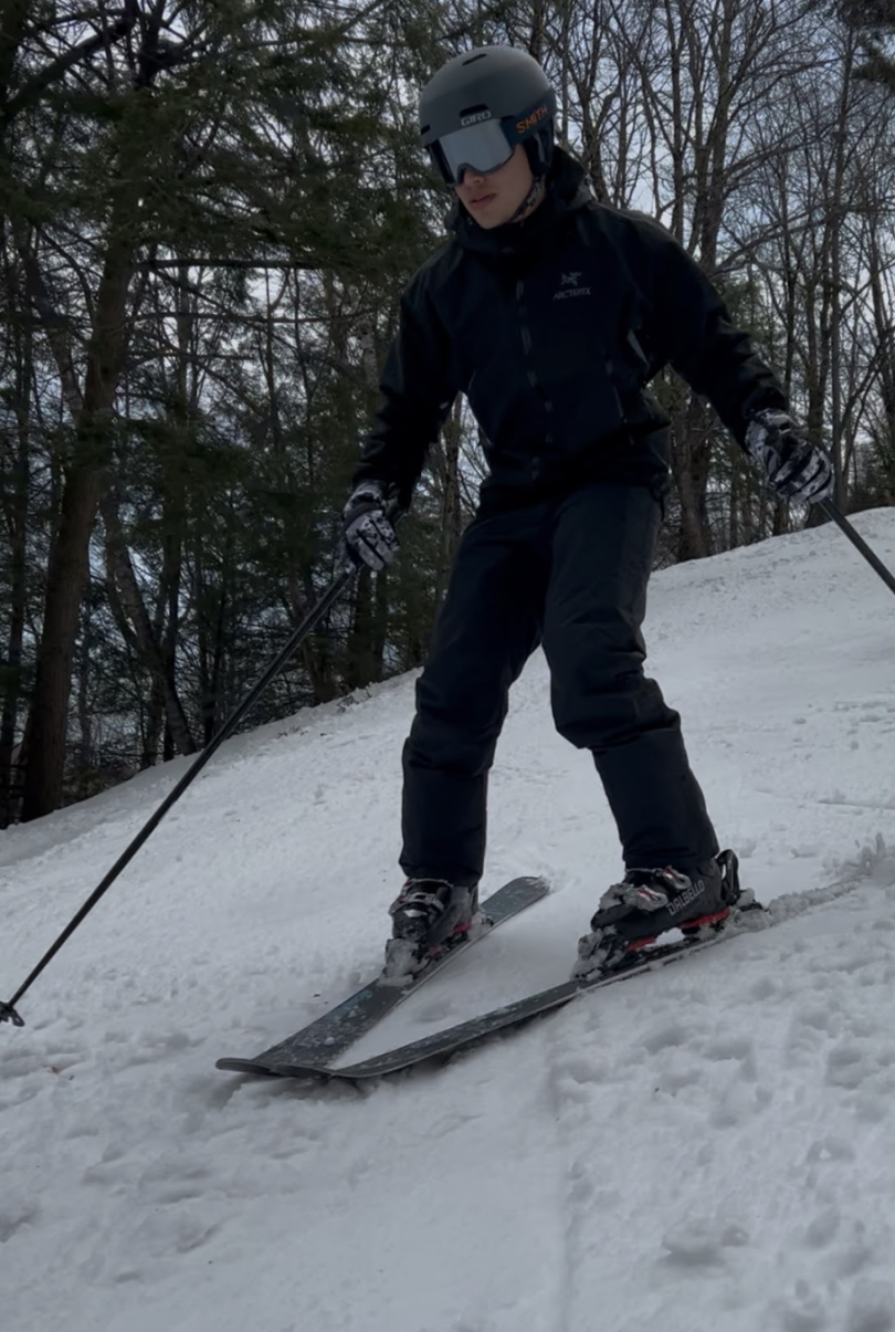 Dartmouth student standing on a slope while wearing skis and holding two ski poles. The student is seen wearing a snow jacket, snow pants, a ski helmet, and ski goggles. The entire ground is covered in white snow, while the sky is clear and blue.