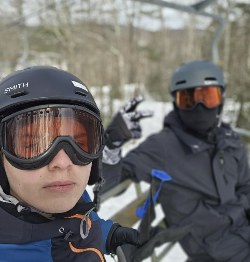 Picture featuring two Dartmouth students wearing ski gear sitting on a chair lift at the Dartmouth Skiway. The image features several trees in the background as well as a clear blue sky.