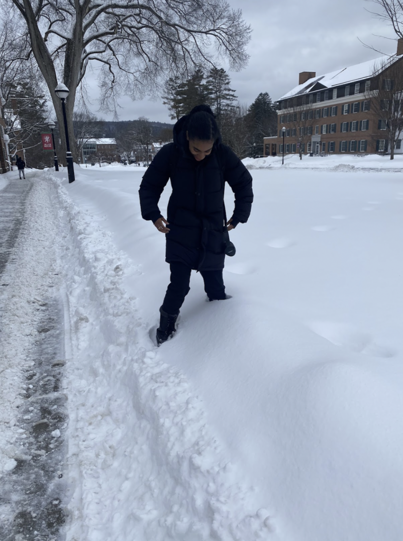 A person wearing a long black coat is standing in deep snow, looking down at their feet. They are partially surrounded by snowdrifts along a path where snow has been cleared. In the background, there are snow-covered buildings and trees under a