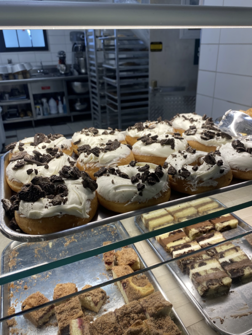 A display of desserts featuring several frosted donuts topped with crushed Oreo cookies and various types of cake slices, including layered cakes and crumb-topped treats.