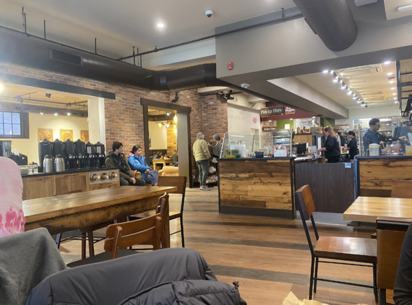 An interior view of a café featuring wooden tables and chairs. The counter is seen in the background with staff preparing drinks, and there are coffee machines and a row of coffee containers along the wall.