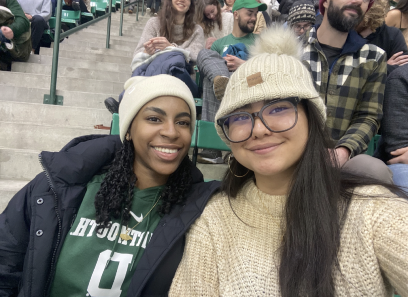 A selfie of two young women smiling while sitting in a hockey stadium. In the background, the stands are filled with people.