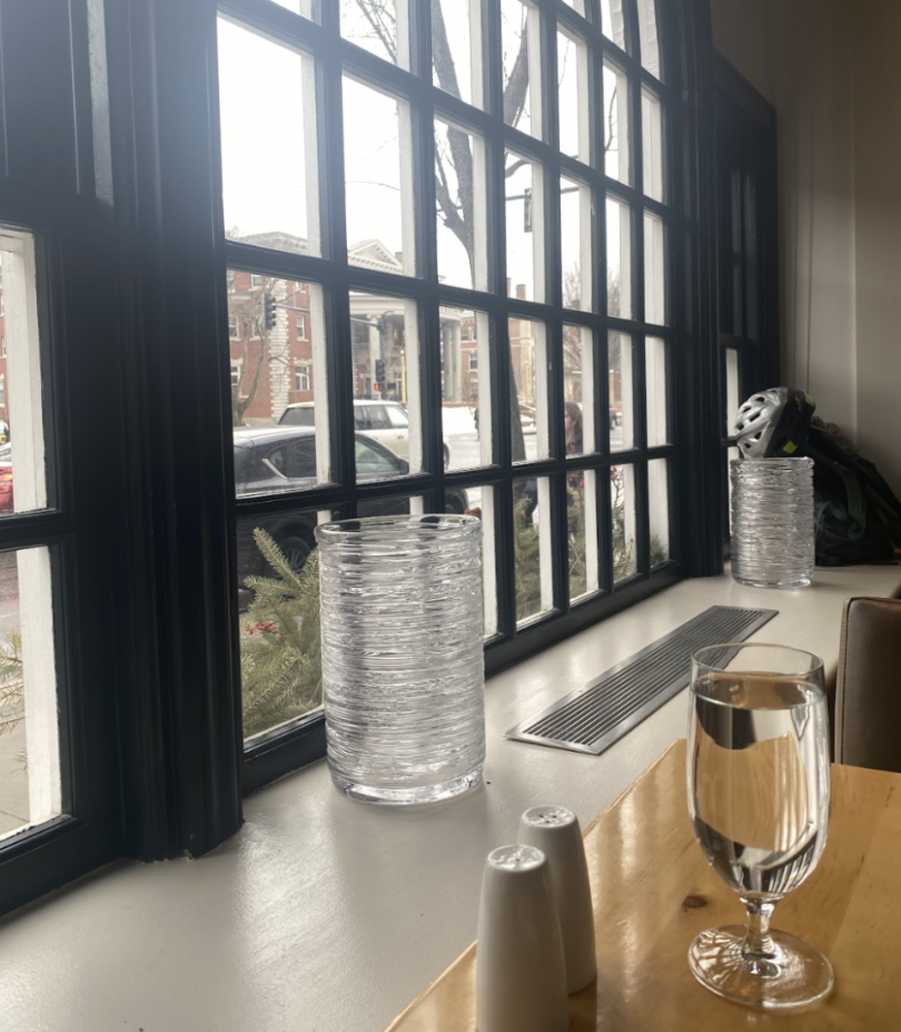 Large arched window at "PINE" restaurant, with a view of the street outside. A wooden table in the foreground holds a glass of water, salt and pepper shakers, and a clear textured vase, with natural light streaming in.