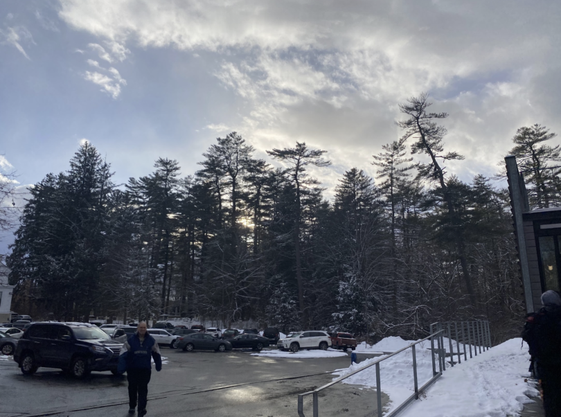 A winter scene with a snowy parking lot surrounded by tall trees. The sky is partly cloudy, with soft sunlight breaking through. Cars are lined up in the lot, and a building is visible in the foreground.