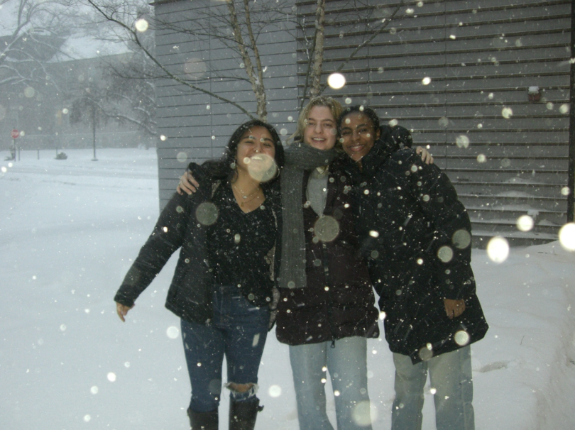 Three friends pose for a photo in a snowy setting, smiling and embracing each other. Snow is falling heavily around them, creating a sparkling atmosphere.