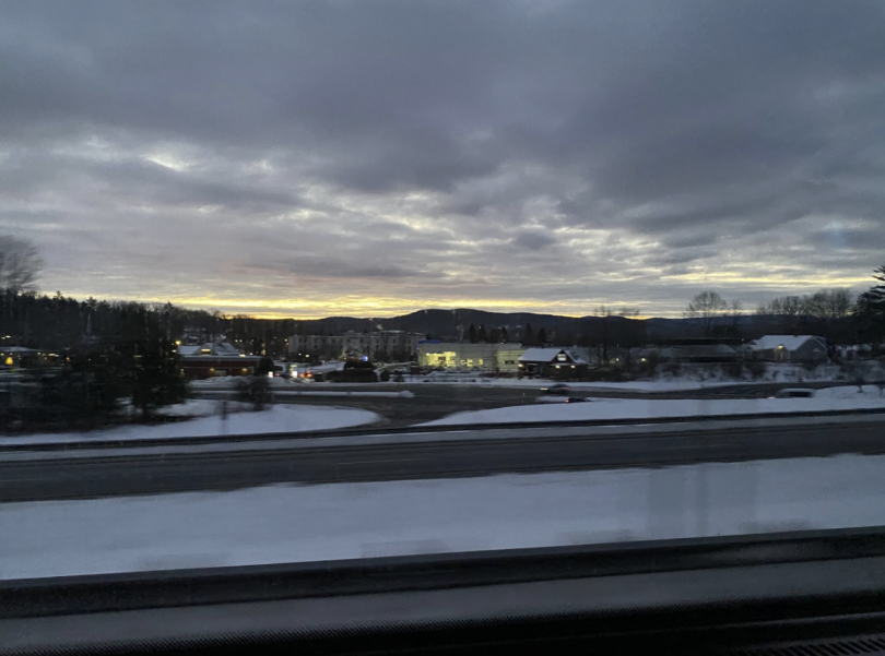 A winter landscape at dawn, showing a highway lined with snow. In the foreground, lights from buildings and cars glisten against the backdrop of a cloudy sky, with hints of sunset colors peeking through the clouds in the distance. Mountains are faintly vi