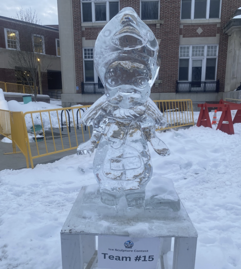 An intricately carved ice sculpture of a sonny angel figure. The sculpture stands on a platform surrounded by a snowy landscape and a backdrop of a building