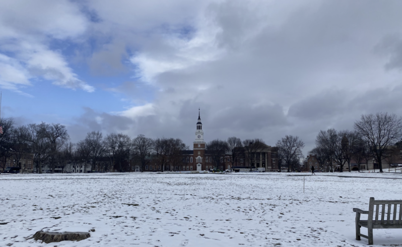 A snowy landscape featuring a large open field with Baker Tower in the background. The sky is partly cloudy, showcasing patches of blue amidst the gray.