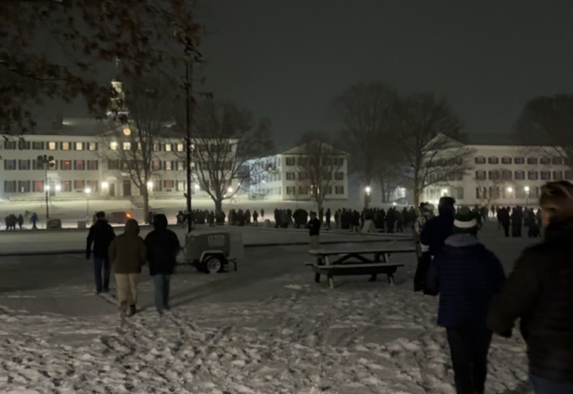 Image featuring Dartmouth Hall and the Green at night. Several people can be seen walking across the green which is covered in a few inches of white snow. Several buildings can be seen in the background with their lights on.