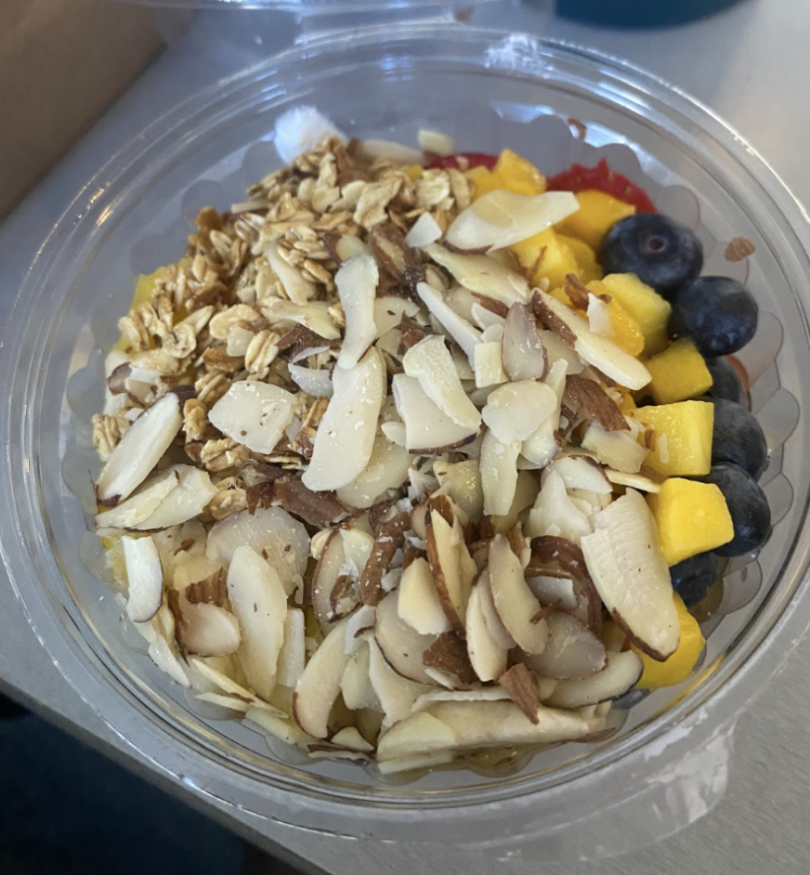 A clear plastic bowl filled with sliced almonds and granola on top of vibrant mango chunks, blueberries, and red berries, resting on a neutral surface.