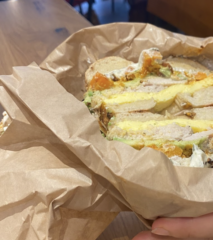  A close-up of a bagel sandwich cut in half, wrapped in brown paper. The sandwich contains eggs, avocado, cream cheese, and possibly smoked salmon or another protein, with a person's hands holding it. The background shows a wooden table and a café setting