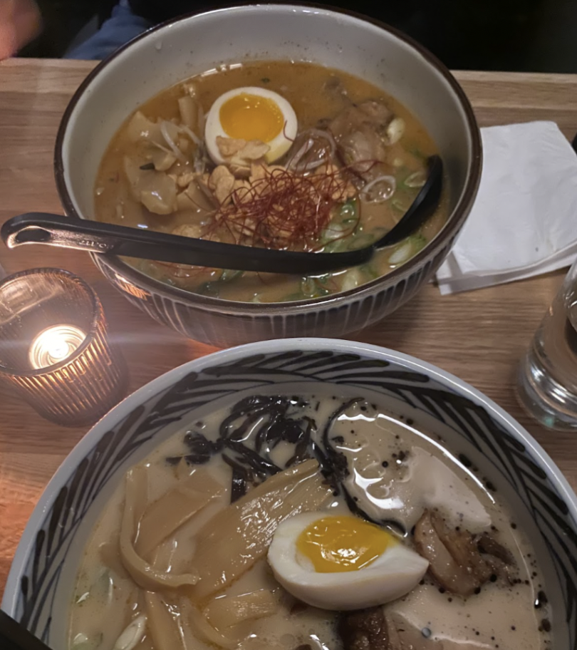 Two bowls of ramen on a wooden table. One bowl contains creamy broth, slices of pork, a soft-boiled egg, bamboo shoots, and dark noodles, while the background bowl has a golden broth with chicken, topped with an egg and green onions.