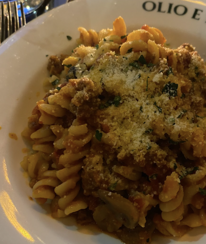 A plate of rotini pasta topped with a rich meat sauce, grated cheese, and sprinkled with fresh herbs. The plate is branded with "OLIO E PIÙ."