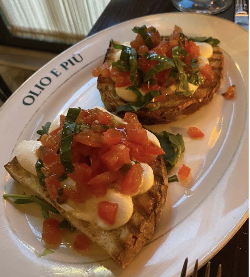 A close-up of a plate from "Olio e Più" with two pieces of bruschetta. The toasted bread is topped with melted mozzarella, diced tomatoes, fresh basil, and a drizzle of olive oil.