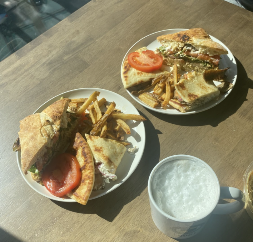 Two plates of Greek-style sandwiches accompanied by crispy French fries, set on a wooden table. The sandwich is cut in half and garnished with tomato slices, while a mug of frothy beverage is nearby, all bathed in natural sunlight.