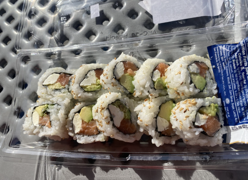 A takeout container with eight sushi rolls filled with salmon, avocado, and cream cheese, topped with sesame seeds. A soy sauce packet rests beside them on a metal table in natural sunlight.