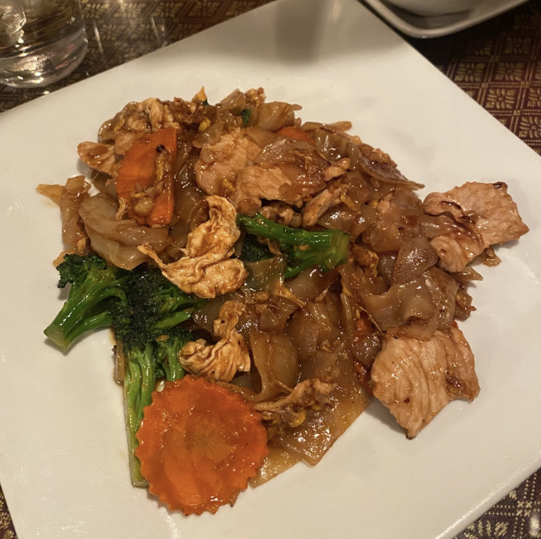 A white square plate holds a serving of stir-fried noodles with chicken, broccoli, carrots, and other vegetables, all glistening with a savory sauce
