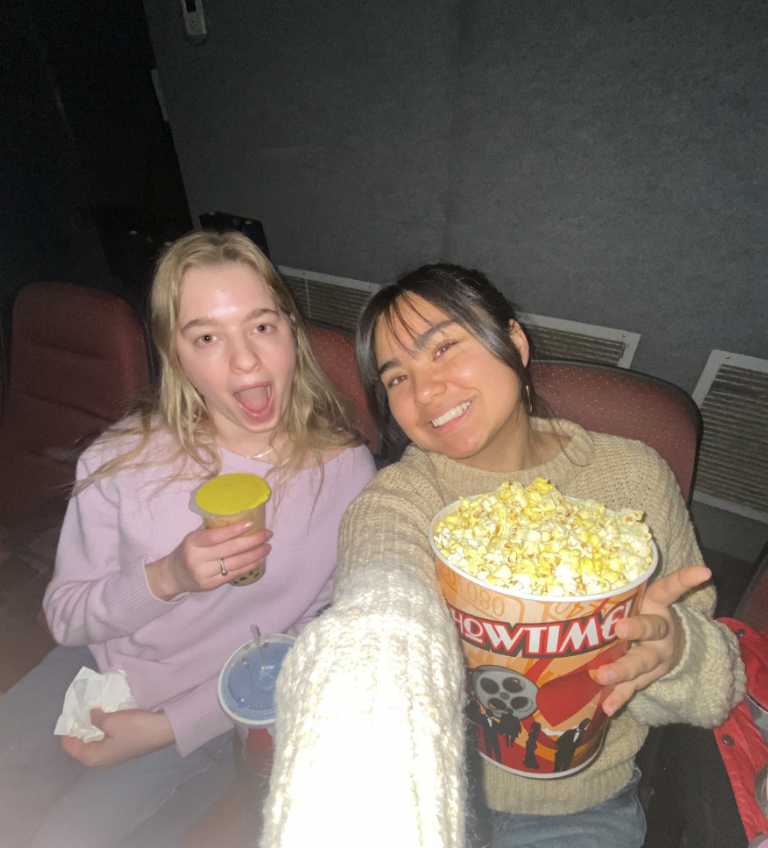Two friends pose for a selfie in a movie theater, smiling and having fun. One holds a large bucket of popcorn, while the other has a small drink.