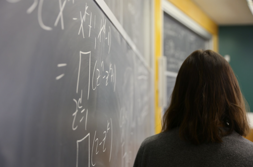The image shows Professor Dougherty-Bliss, who has shoulder-length brown hair and is wearing a dark sweater, standing in front of a large blackboard filled with mathematical equations written in white chalk. The perspective is from behind, with the focus