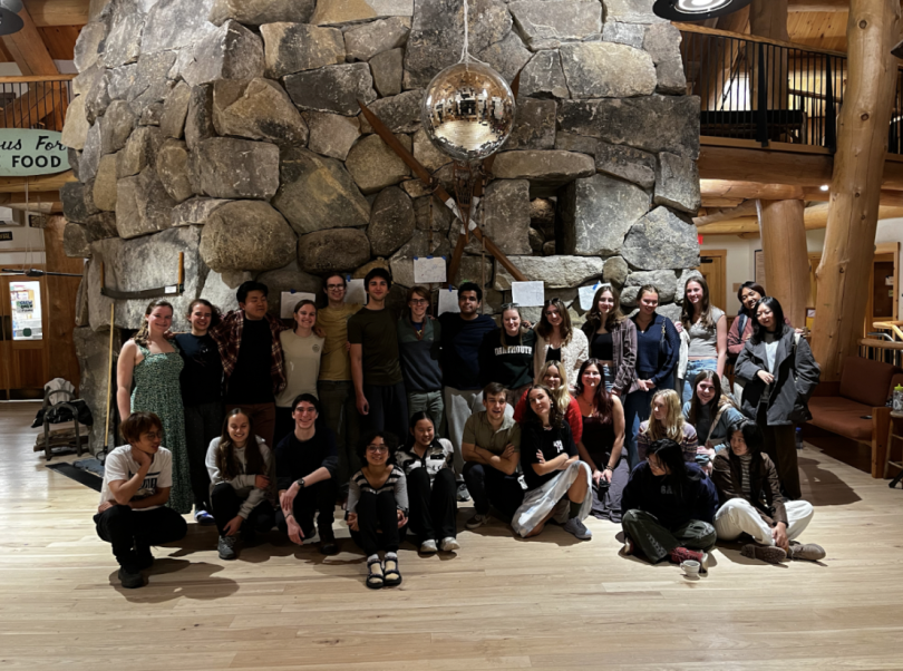 This photo captures a big chunk of CnT members gathered inside a rustic lodge, posing in front of a massive stone fireplace adorned with a disco ball. Everyone is having fun and the people are spread around the whole picture, with people standing, sitting