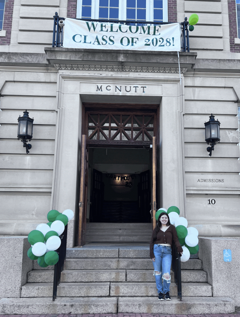 A girl in blue jeans and a brown sweater stands on the steps of a stone building. The doorway reads "McNutt" and a white banner hangs overhead, which reads "Welcome Class of 2028!"