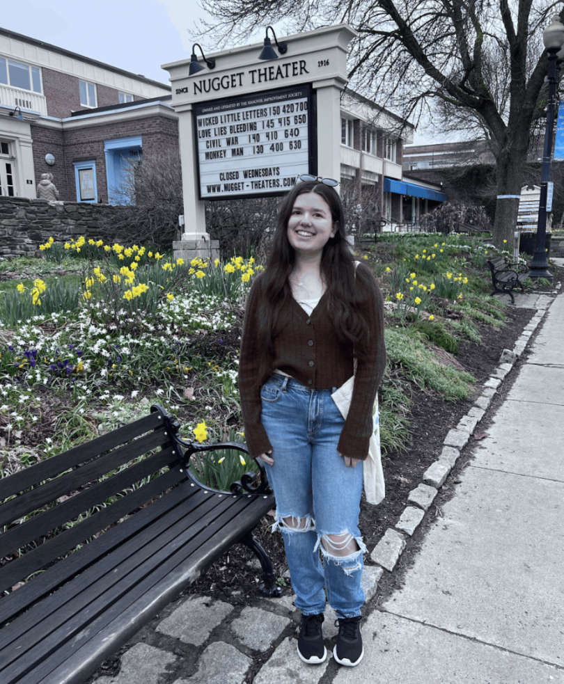 A girl in blue jeans and a brown sweater stands in front of a movie theater marquee.