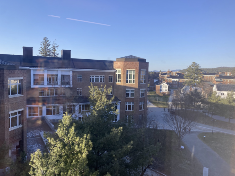 View of a red-brick academic building surrounded by trees, with clear blue skies and more campus buildings in the distance.