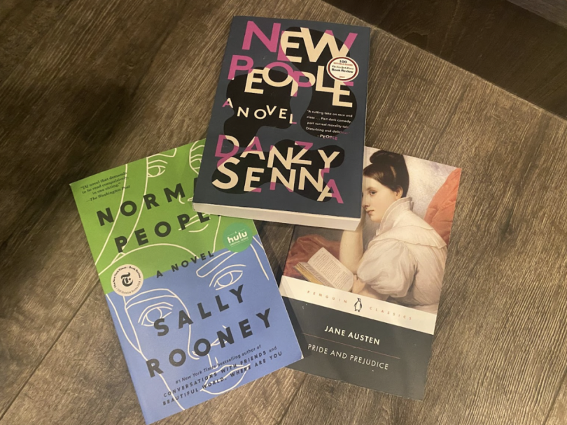 Three novels — New People by Danzy Senna, Normal People by Sally Rooney, and Pride and Prejudice by Jane Austen — laid out on a wooden table.