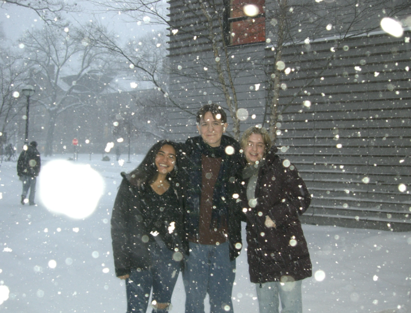 Three friends smiling and posing outside in heavy snowfall near a building, dressed in winter clothing.