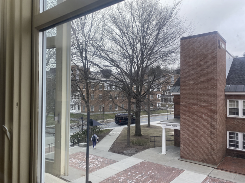 View from a window overlooking a brick campus building labeled "Raven House," with bare trees, overcast skies, and a few people walking along the path.