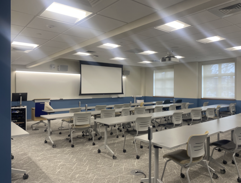 A bright, modern classroom with rolling chairs, long tables, a projector screen, and large windows along one wall.