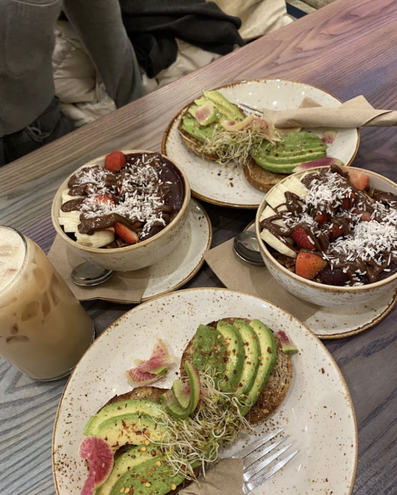 A colorful brunch spread featuring avocado toast topped with sprouts and radish, acai bowls with fruit and coconut, and an iced latte on a rustic wooden table.