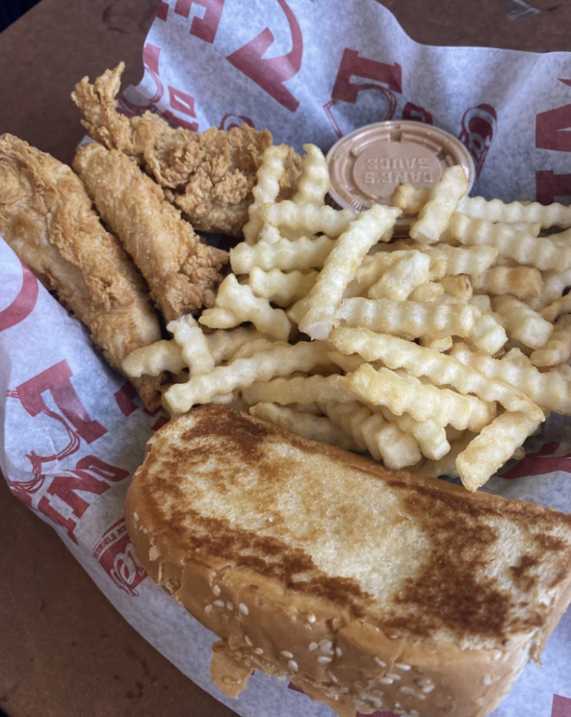 A Raising Cane's meal featuring crispy chicken tenders, crinkle-cut fries, Texas toast, and a container of Cane's Sauce on branded paper.