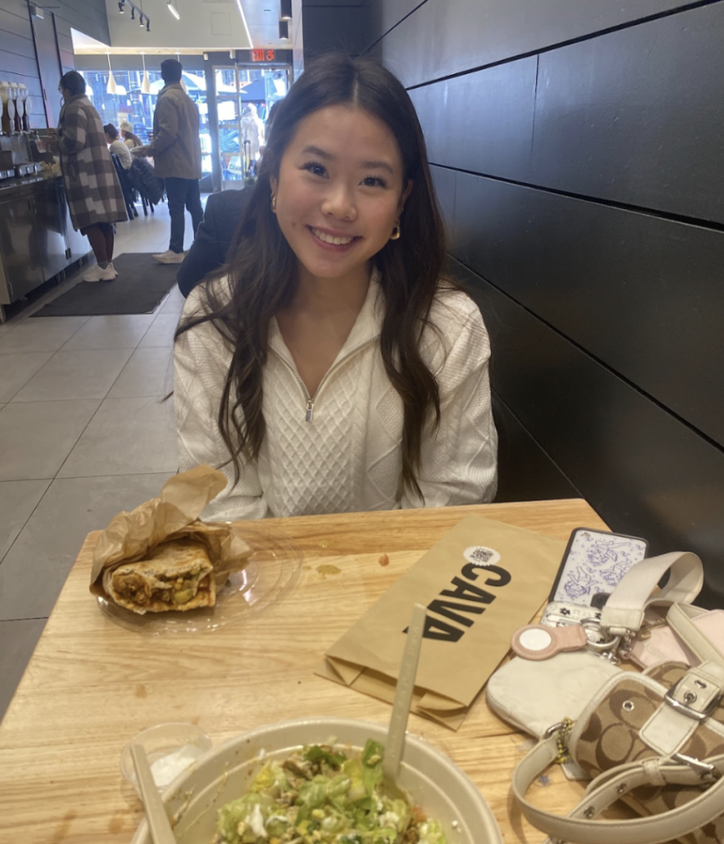 A young woman in a white sweater smiles at a table at a Cava restaurant, with a wrap and salad